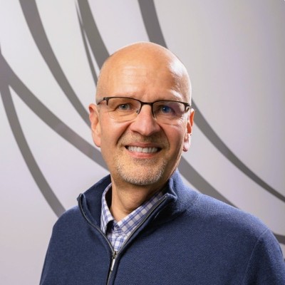 Head-and-shoulders portrait of a smiling, bald man with glasses wearing a blue zip-up sweater, in front of an abstract circular backdrop.